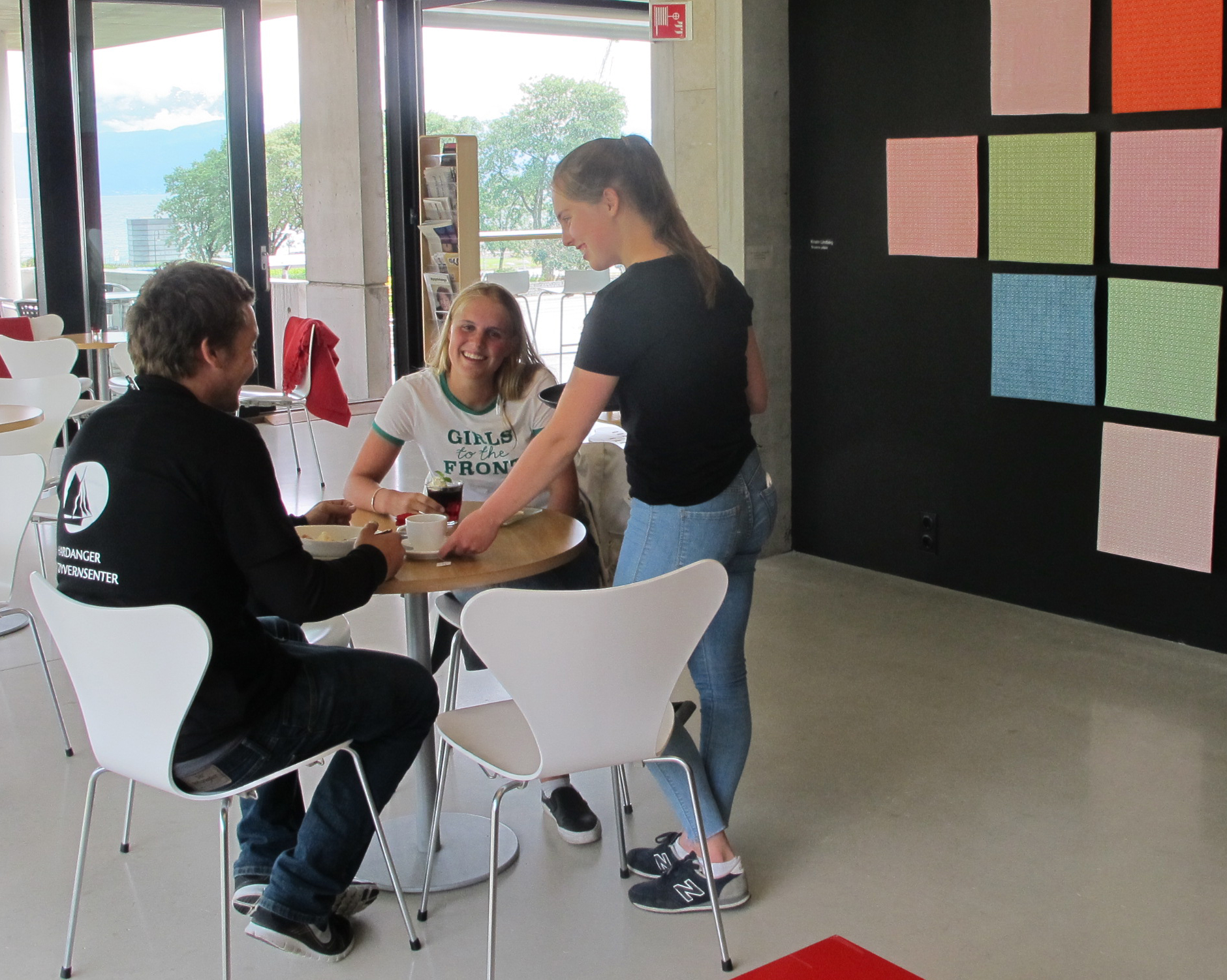 To mennesker ved et kafebord i et lyst rom. med kunst på veggene. En annen person står ved bordet.