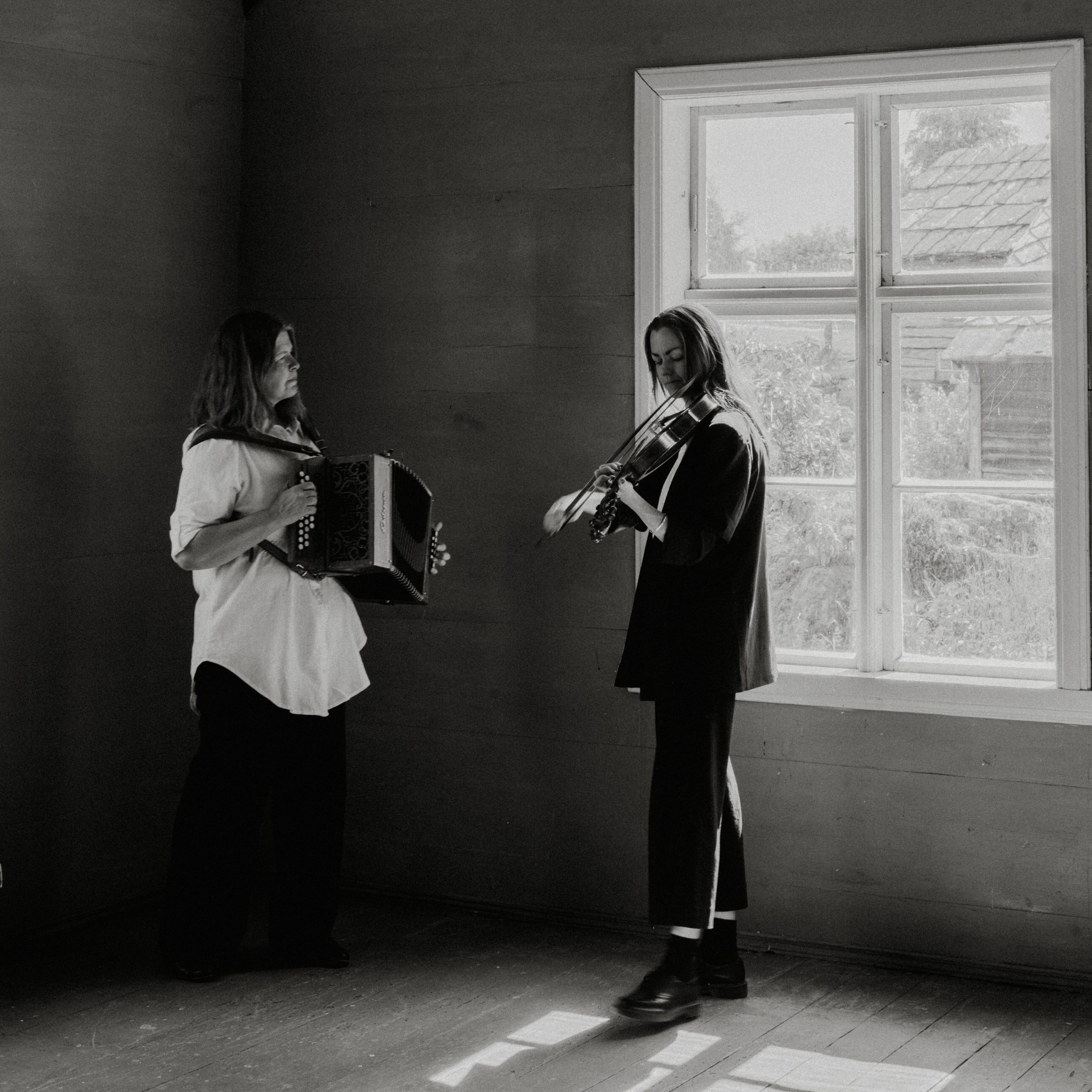 to damer står i et rom med eit stort vindu, den ene spelar toradar den adre hardingfele