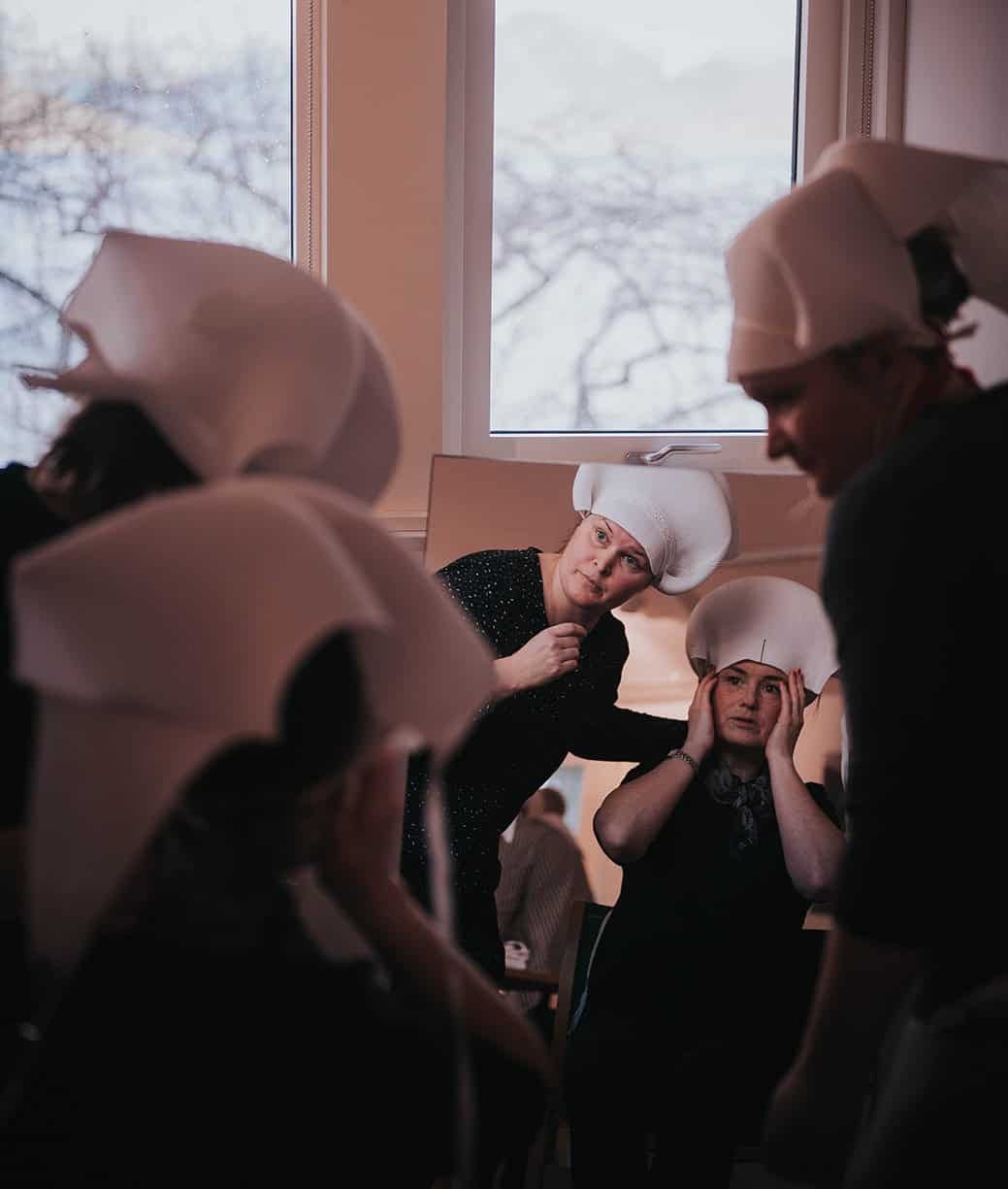 To kvinner med bunadskaut på hodet ser seg selv i speilet og ser om skautet sitter riktig