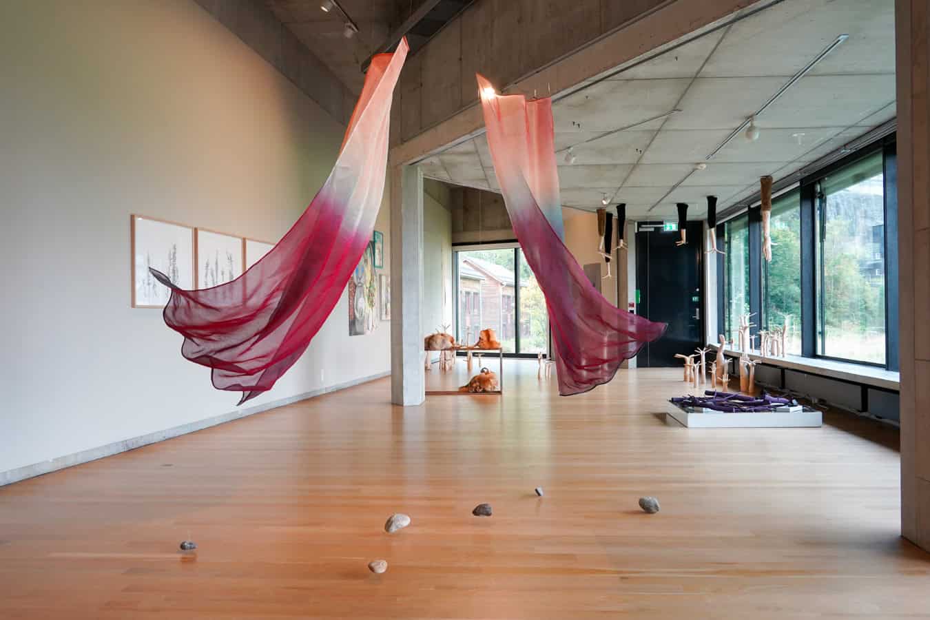 Vestlandsutstillingen 2025 Utstillingsrom i Kunsthuset Kabuso med hengande tekstilar i raudtonar og treskulpturar i rommet, med store vindauge mot hagen utanfor