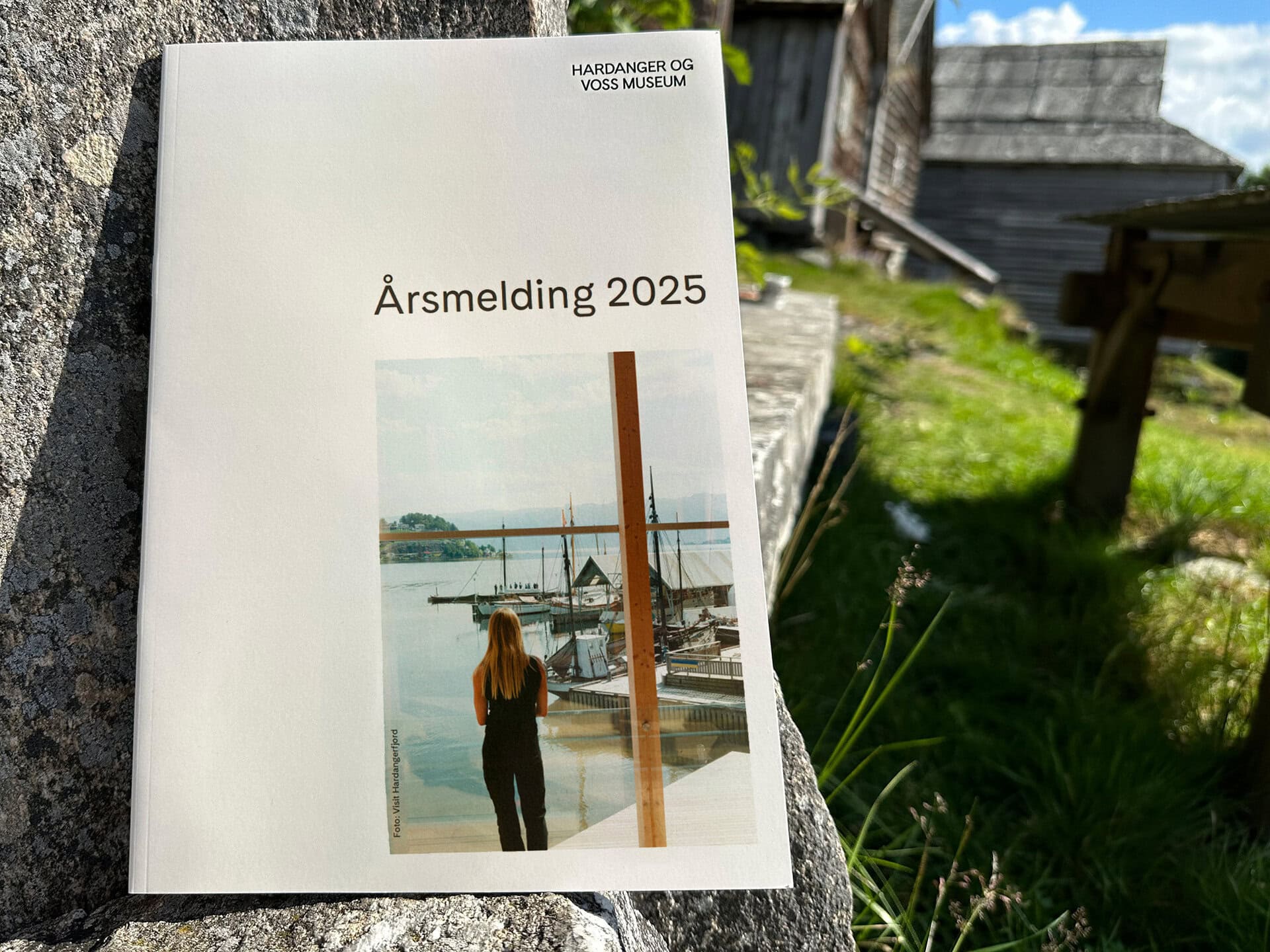 ÅrsmeldinG HVM 2025 illustrasjon Årsmeldingen 2025 til Hardanger og Voss museum med bilde av en person som står og ser utover en brygge med båter. Årsmeldingen står på en steinplate og har gamle bygg bak seg.